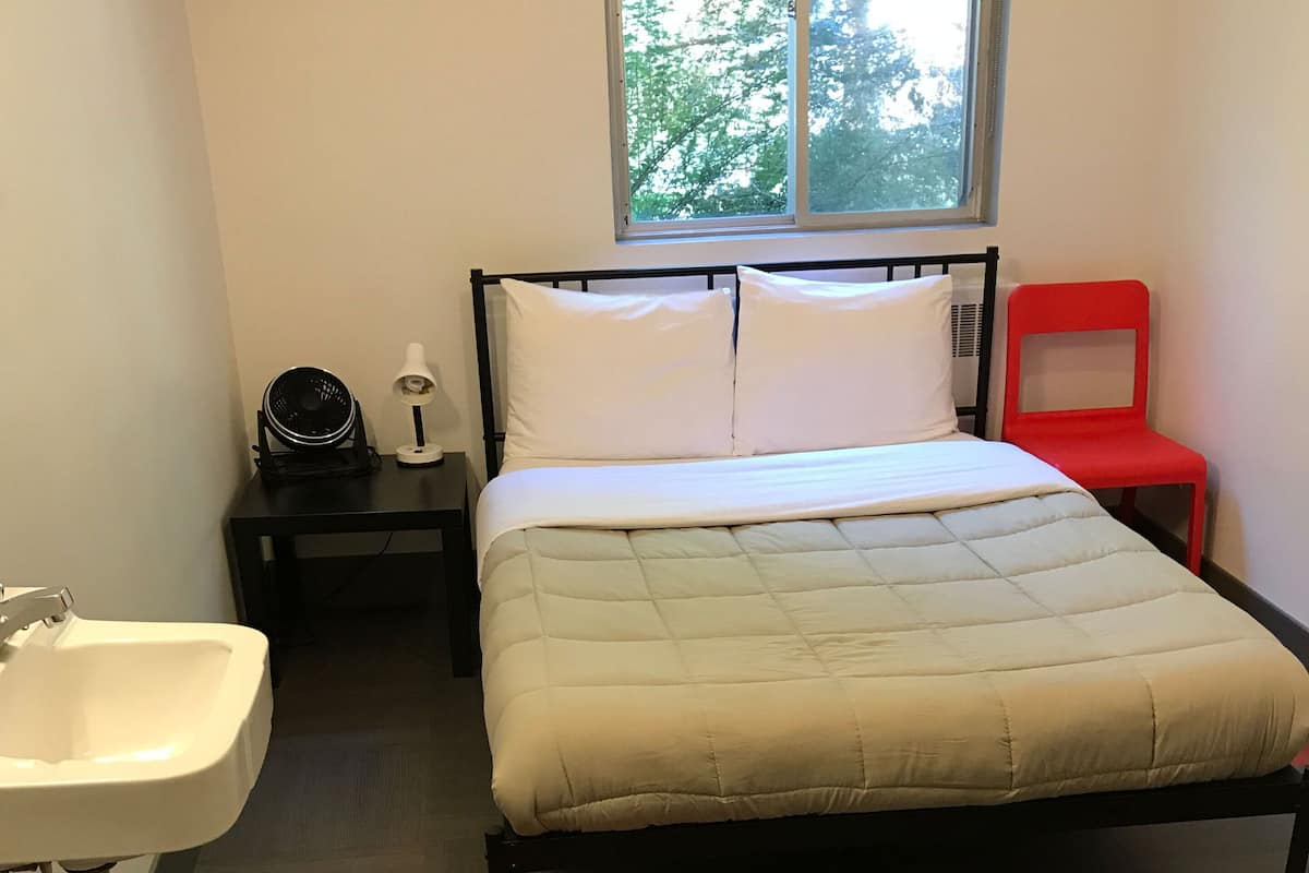 A bedroom with a bed, a nightstand, a red chair, a window, and a sink.