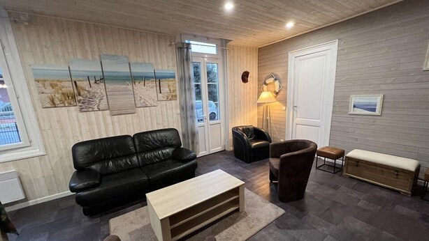 Uma sala de estar com sofá de couro preto, mesa de centro de madeira, poltrona marrom e um banco. Há uma janela grande com vista para o exterior, uma porta e uma parede com um mural de praia.