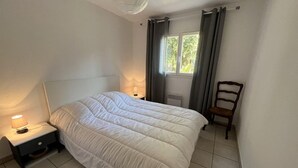 Ein Schlafzimmer mit einem Bett, einem Nachttisch mit Lampe, einem Sessel und einem Fenster mit Vorhängen.