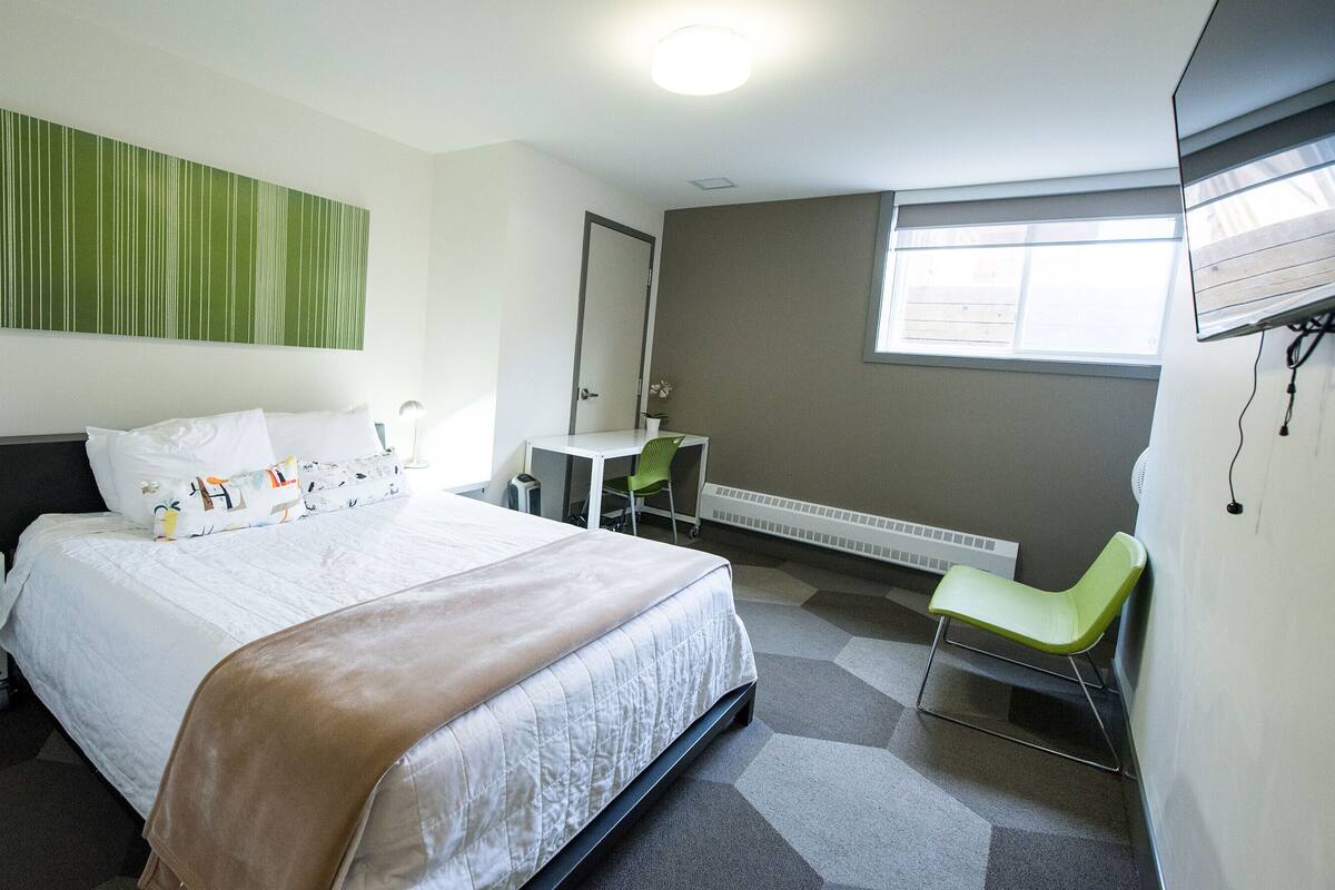 A hotel room with a bed, a desk, a chair, a heater, and a window.