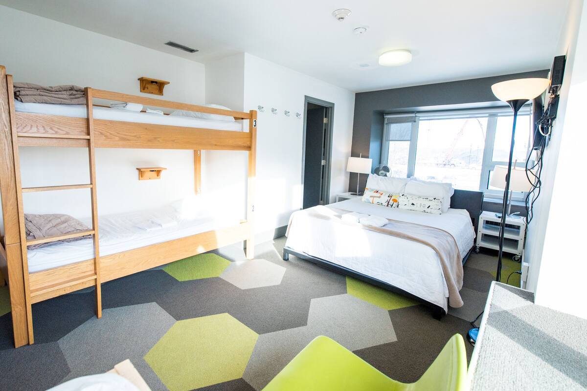 A hotel room with a bunk bed, a single bed, a desk with a chair, a lamp, and a window with a view of the outside.