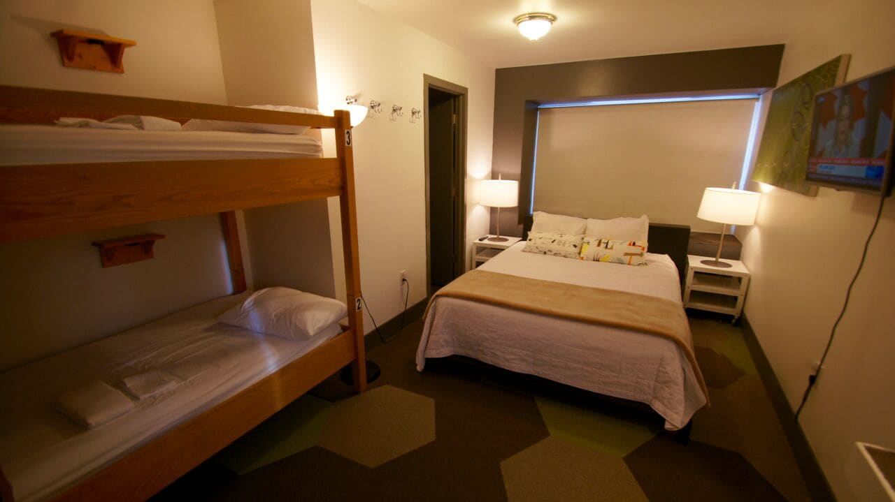Ein Hotelzimmer mit Etagenbett, einem Einzelbett, Nachttischlampen, einem Nachttisch und einem an der Wand befestigten Fernseher.