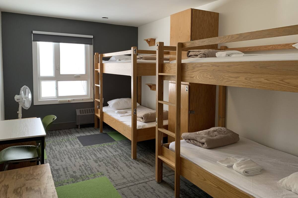 A dormitory room with bunk beds, a desk, a chair, a fan, and a window.
