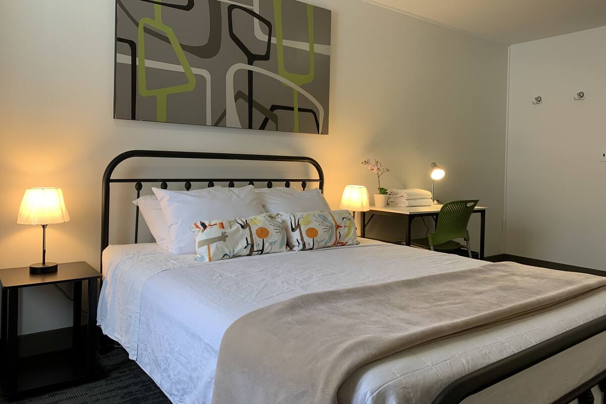 A neatly made bed with a white comforter and decorative pillows, a bedside table with a lamp, and a wall-mounted abstract art piece.