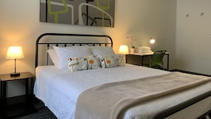 Una cama bien hecha con una colcha blanca y almohadones decorativos, una mesita de noche con lámpara, y un cuadro abstracto colgado en la pared.