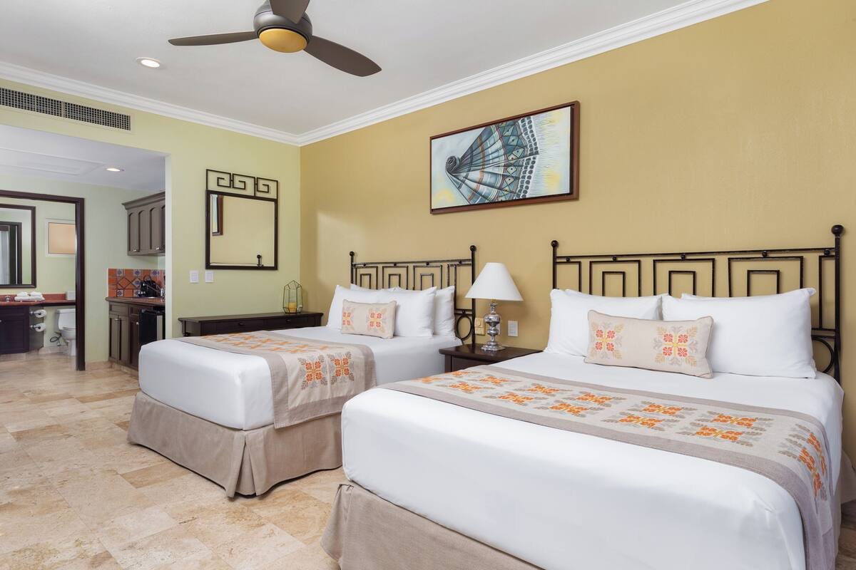 Une chambre d’hôtel avec deux lits, un ventilateur de plafond, un tableau, un miroir et une salle de bain visible à travers une porte ouverte.