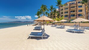 A beach with sun loungers and umbrellas, a building with balconies, and palm trees. - Villa del Palmar Flamingos Beach Resort & Spa (Nuevo Nayarit)