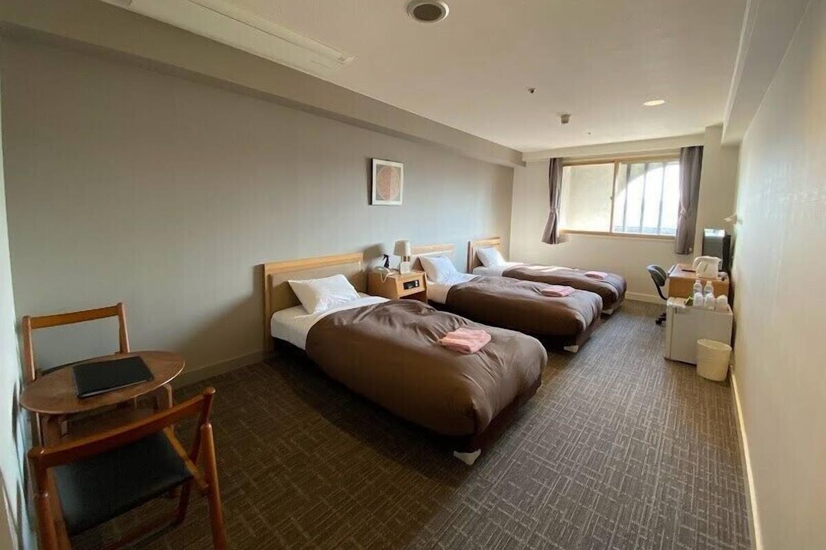 Une chambre d’hôtel avec deux lits, un bureau, une chaise, une fenêtre et une petite table avec une lampe.
