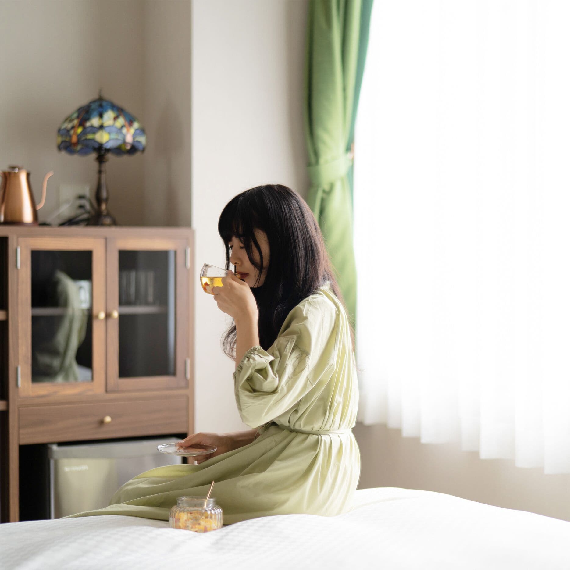 A person sitting on a bed in a room with a wooden cabinet and a lamp.