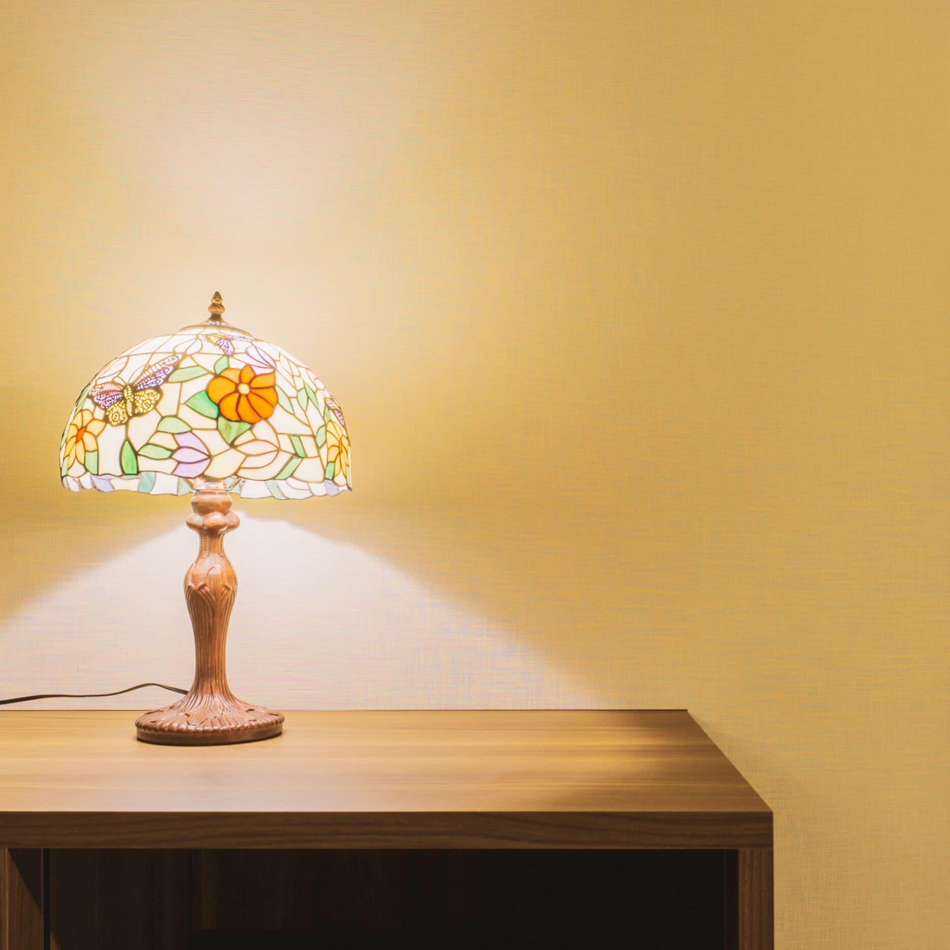 A table lamp with a stained glass shade on a wooden table against a plain wall.