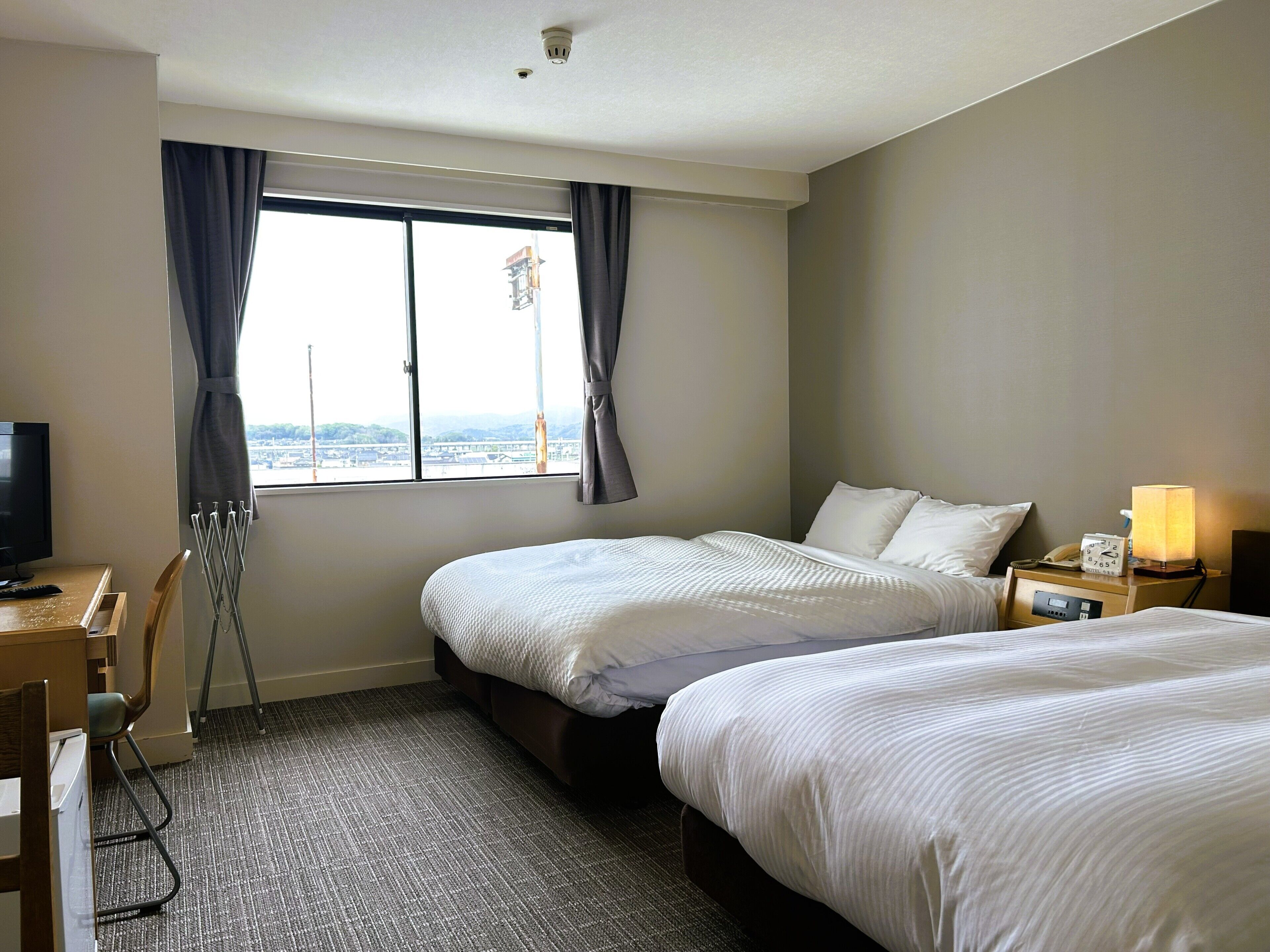 A hotel room with two beds, a desk with a chair, a TV, a window with curtains, and a nightstand with a lamp.