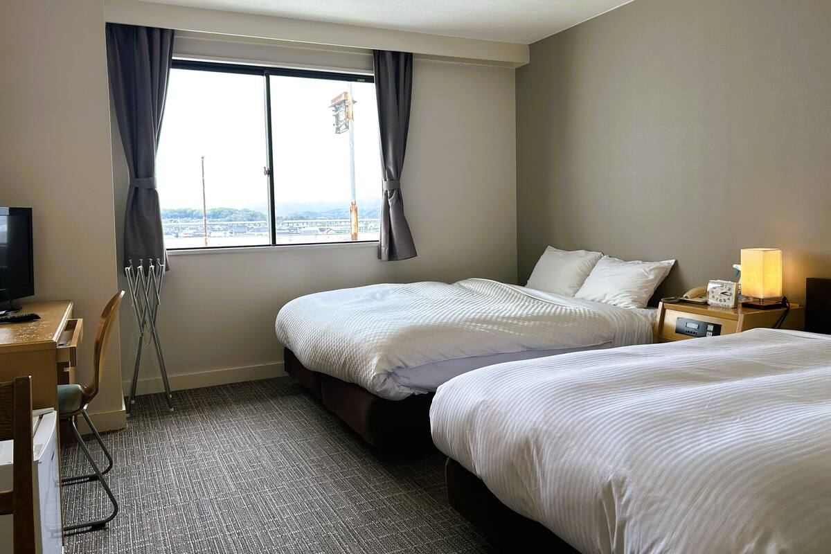 Une chambre d’hôtel avec deux lits, un bureau avec une chaise, une télévision, une fenêtre avec des rideaux et une table de chevet avec une lampe.