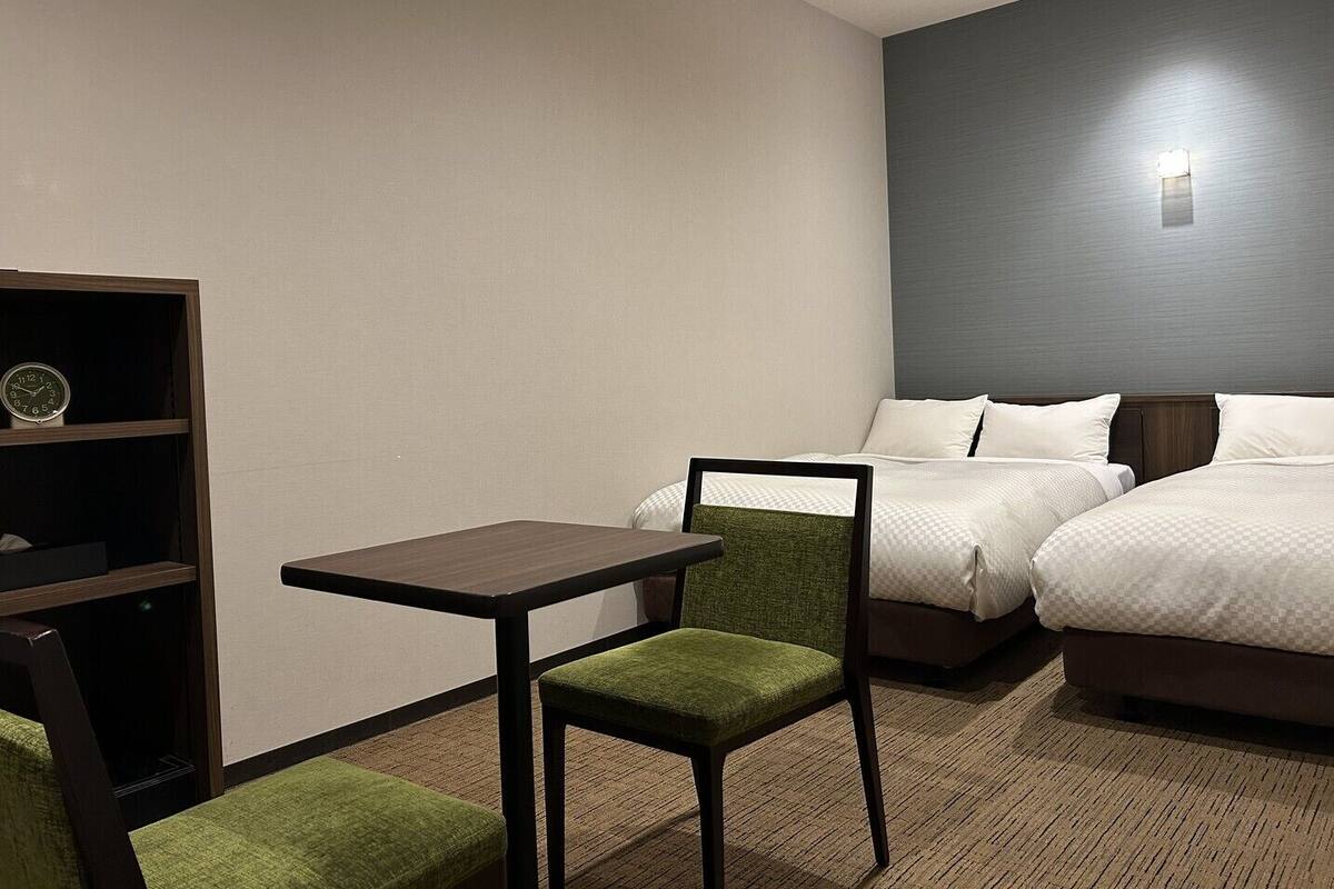 Une chambre d’hôtel avec un lit, un bureau, une chaise et une étagère sur laquelle se trouve une horloge.