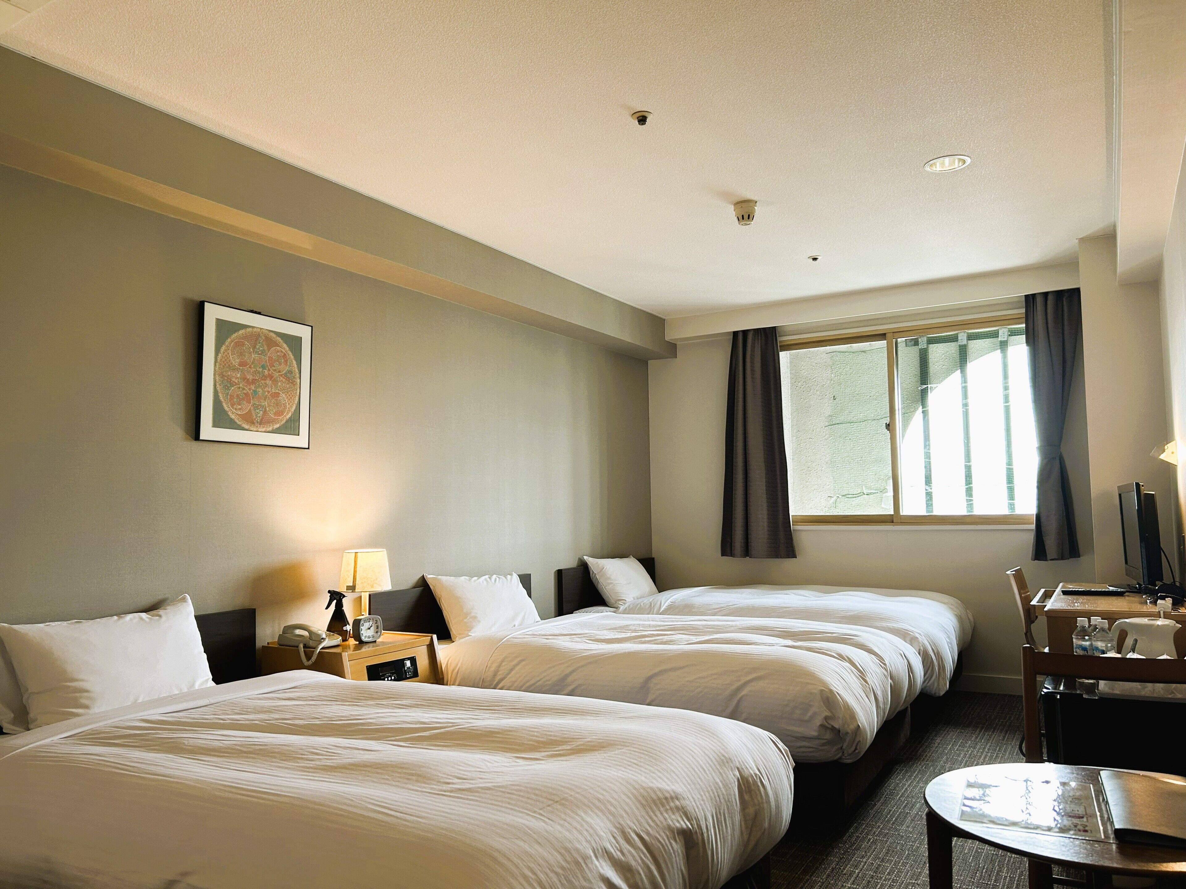A hotel room with two beds, a desk, a chair, a window with curtains, a wall-mounted picture, and a ceiling light.