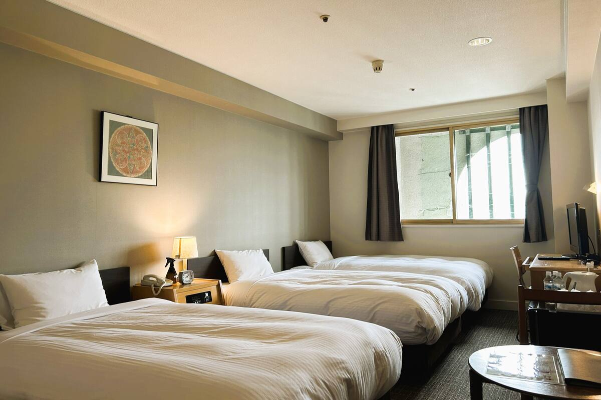 Une chambre d’hôtel avec deux lits, un bureau, une chaise, une fenêtre avec des rideaux, un tableau accroché au mur et un luminaire au plafond.