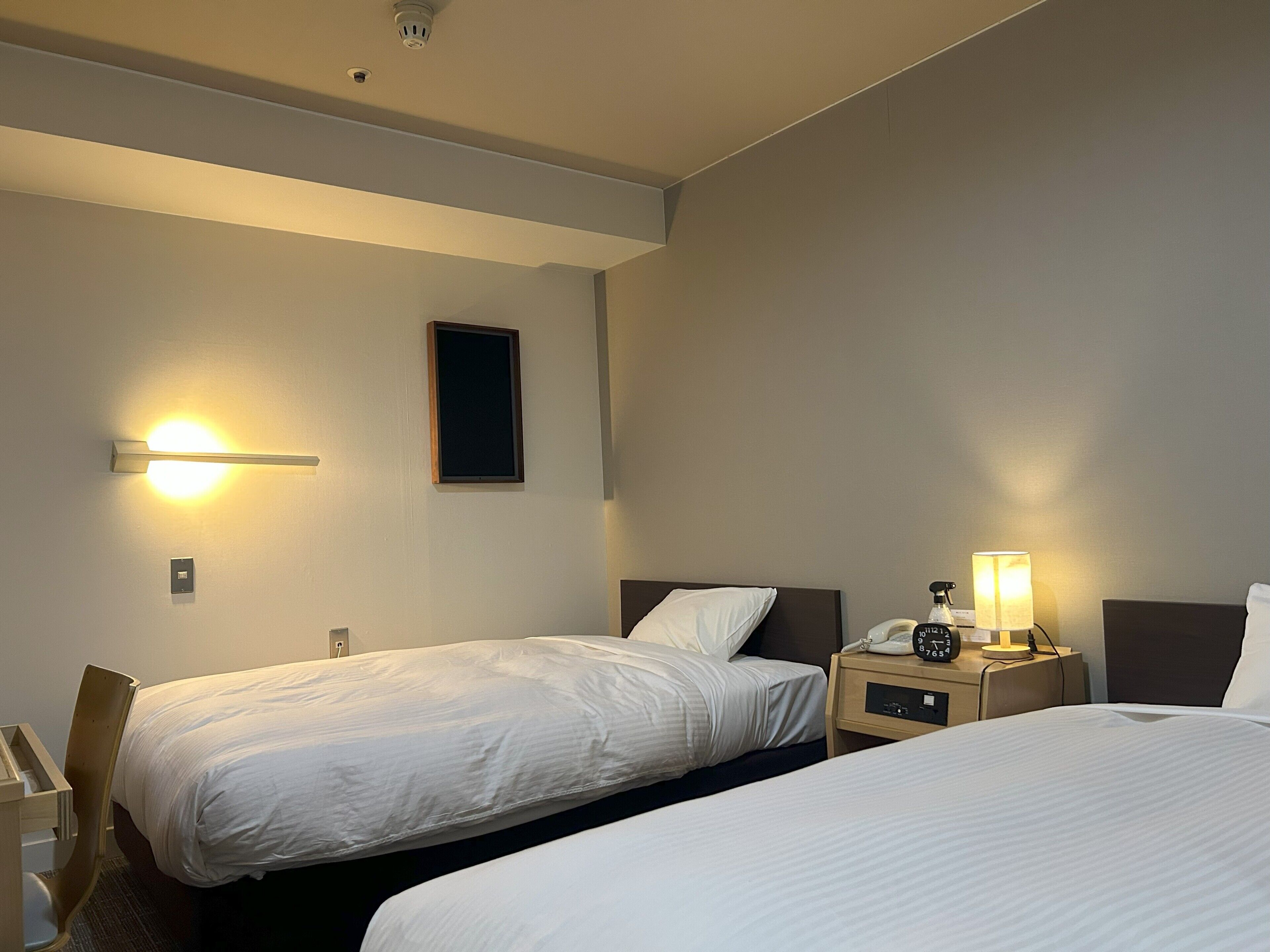 A hotel room with two beds, a nightstand with a lamp, a phone, and a wall-mounted light fixture.