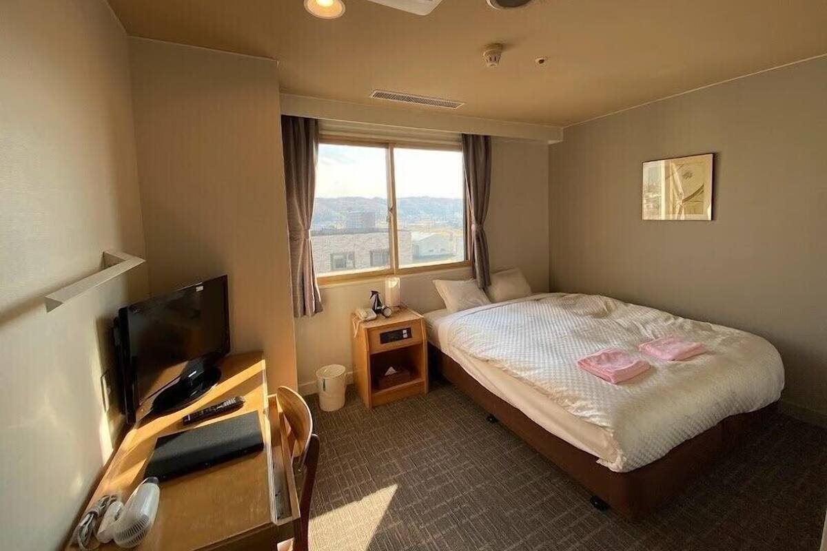 Une chambre d’hôtel comprenant un lit, un bureau avec une télévision, une chaise, une fenêtre donnant sur la ville et une table de chevet avec une lampe.