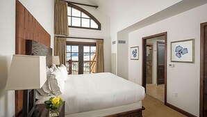 A hotel room with a large bed, a desk with a lamp, a window with curtains, and a door leading to another room.