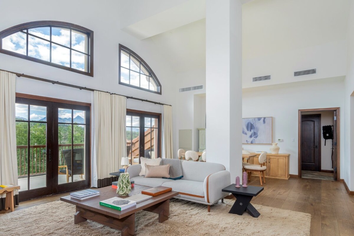A spacious living room with a large sofa, a coffee table, and a view of mountains through the windows.