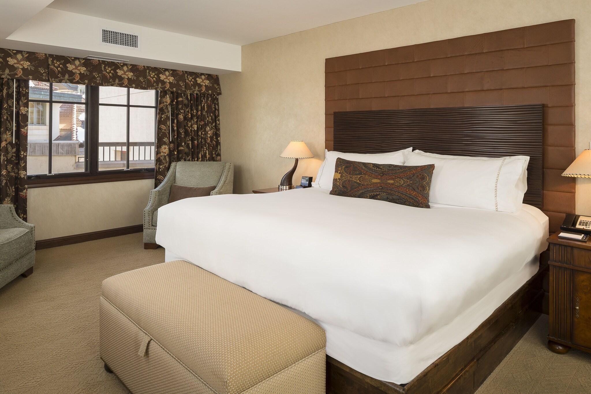 Une chambre d’hôtel avec un grand lit, deux tables de chevet, une chaise, une fenêtre avec des rideaux et une vue sur les bâtiments environnants.
