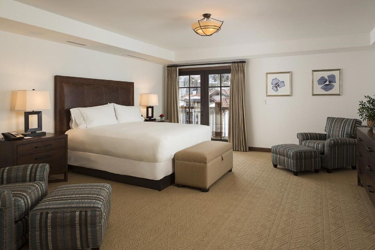 A hotel room with a large bed, two armchairs, a small table, a lamp, a dresser, and a window with curtains.