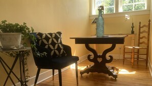 A room with a wooden floor, a black chair with a geometric cushion, a small table with a vase, and a window with natural light. - Sunny Blue Bungalow near Nashville in Historic Downtown Murfreesboro (Murfreesboro)
