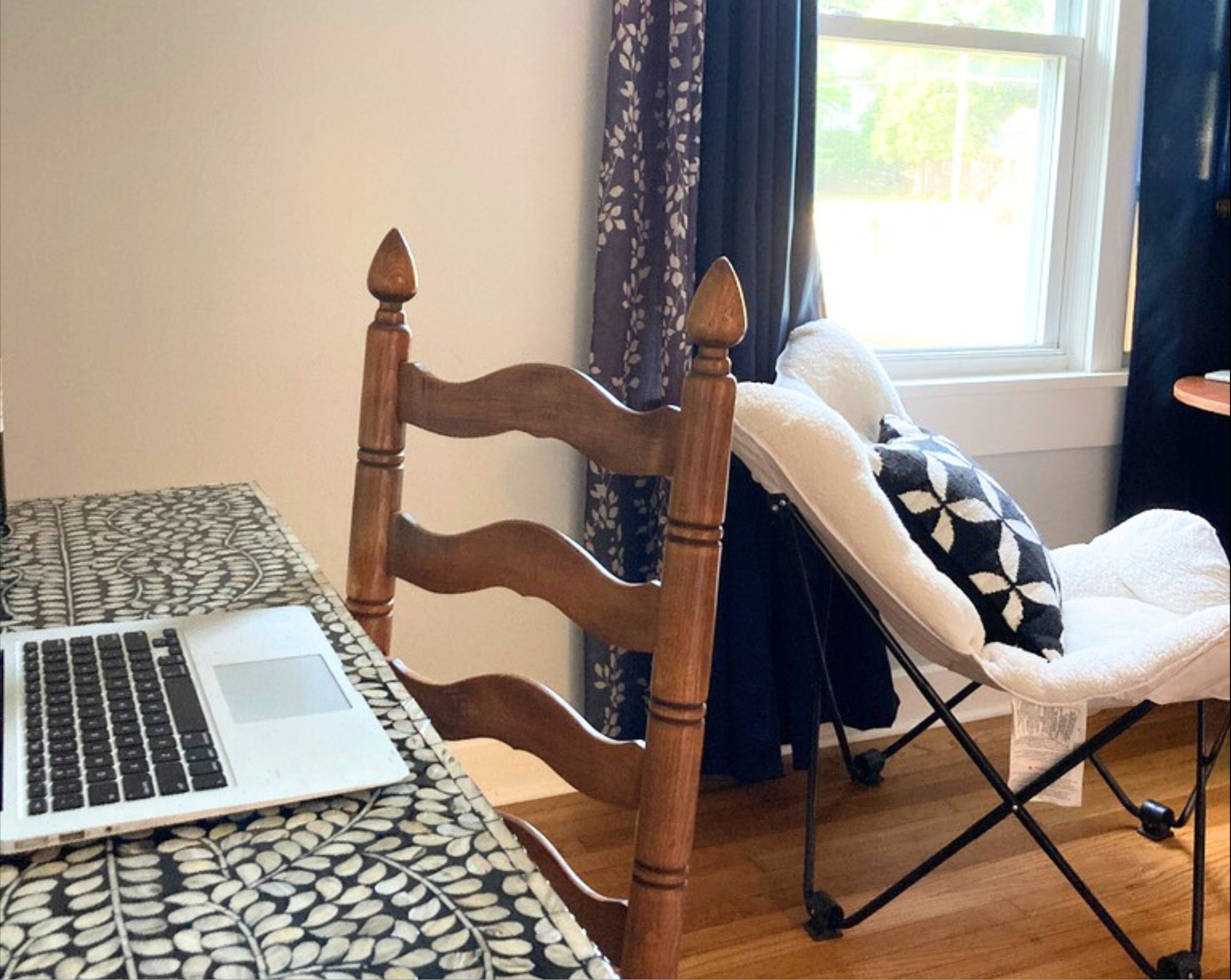 A wooden chair with a cushioned backrest, a laptop on a patterned table, and a window with curtains.
