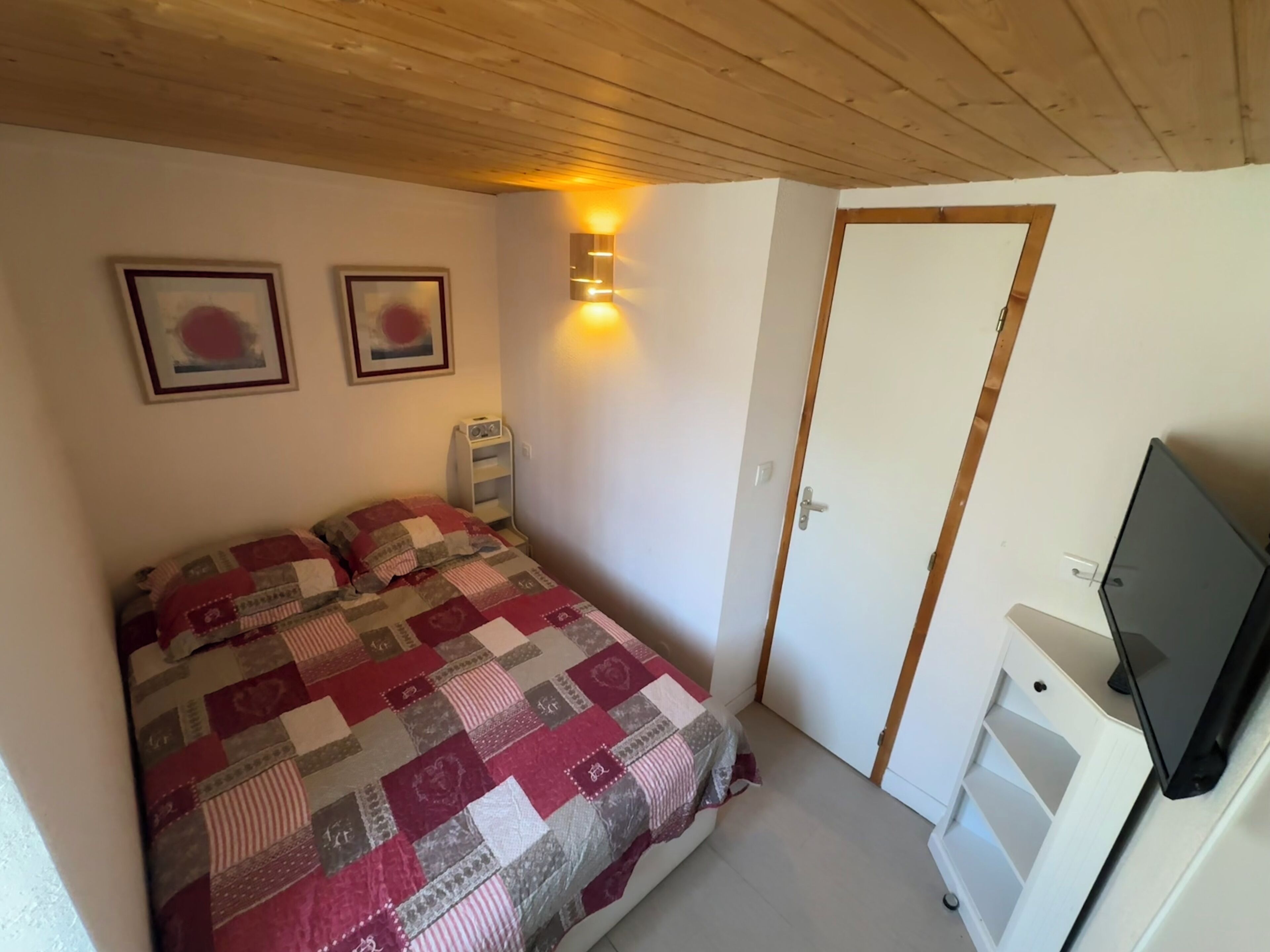 Ein Schlafzimmer mit einem Bett, zwei gerahmten Bildern, einem Nachttischradio, einer Tür und einem Fernseher auf einem Schrank.