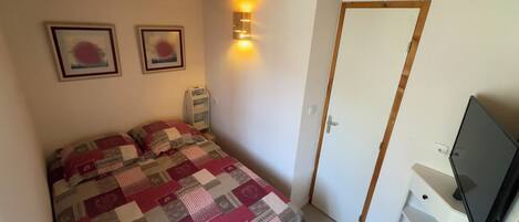 Ein Schlafzimmer mit einem Bett, zwei gerahmten Bildern, einem Nachttischradio, einer Tür und einem Fernseher auf einem Schrank.