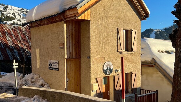 Un chalet rustique avec une porte en bois et des volets aux fenêtres, entouré d’un paysage enneigé et dominé par des montagnes en arrière-plan.