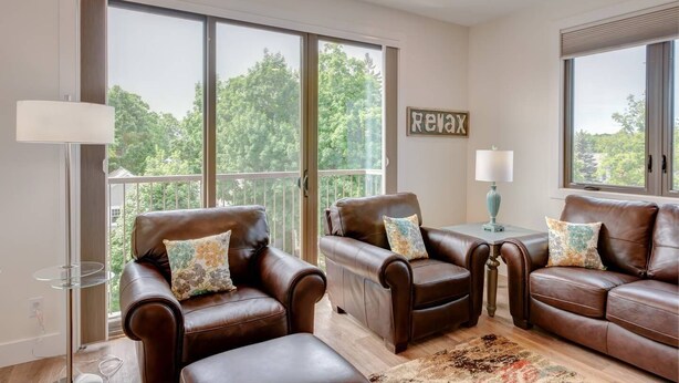 A spacious living room with large windows, two leather sofas with patterned cushions, a small table with a lamp, and a 'Relax' sign on the wall. - Capri TC: Charming Downtown TC Oasis (Traverse City)
