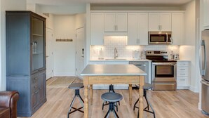 A modern kitchen with white cabinets, stainless steel appliances, and a wooden island with stools. - Capri TC: Charming Downtown TC Oasis (Traverse City)