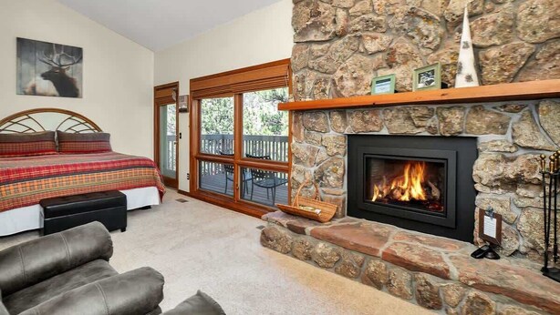 A bedroom with a stone fireplace, a bed with a plaid bedspread, and a view of the outdoors through a glass door. - Deer Mountain Retreat near RMNP! #3206 (Estes Park)
