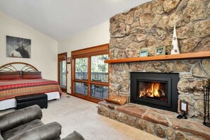 A bedroom with a stone fireplace, a bed with a plaid bedspread, and a view of the outdoors through a glass door.