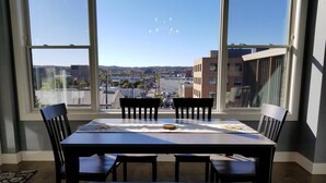 Un comedor con mesa y sillas, ventanas amplias con vistas a edificios y un paisaje lejano.