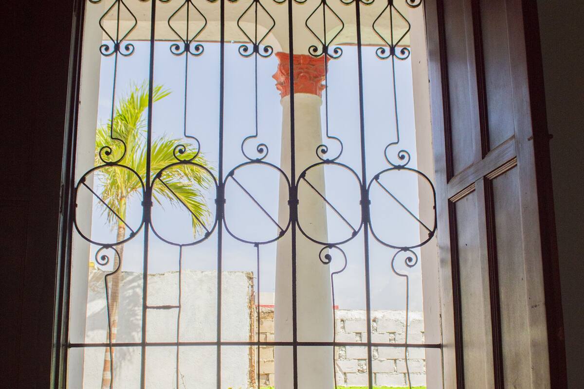 A room with a bed, a window with decorative metal bars, and a view of a palm tree and a building outside.