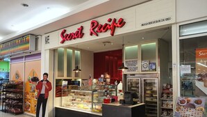 Una cafetería llamada 'Secret Recipe' con un expositor de vidrio lleno de pasteles y un letrero que indica el horario de atención.