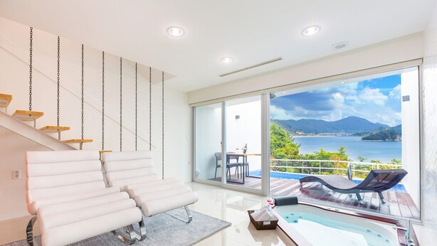 A modern interior with a white sofa, a glass-enclosed balcony with a view of a lake and mountains, and a hot tub. - Geoje Star Marine Pool Villa Pension - V (Geoje)