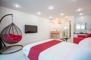 A modern hotel room with a hanging chair, a bed with red bedding, a TV, a kitchenette, and a bathroom with a mirror.