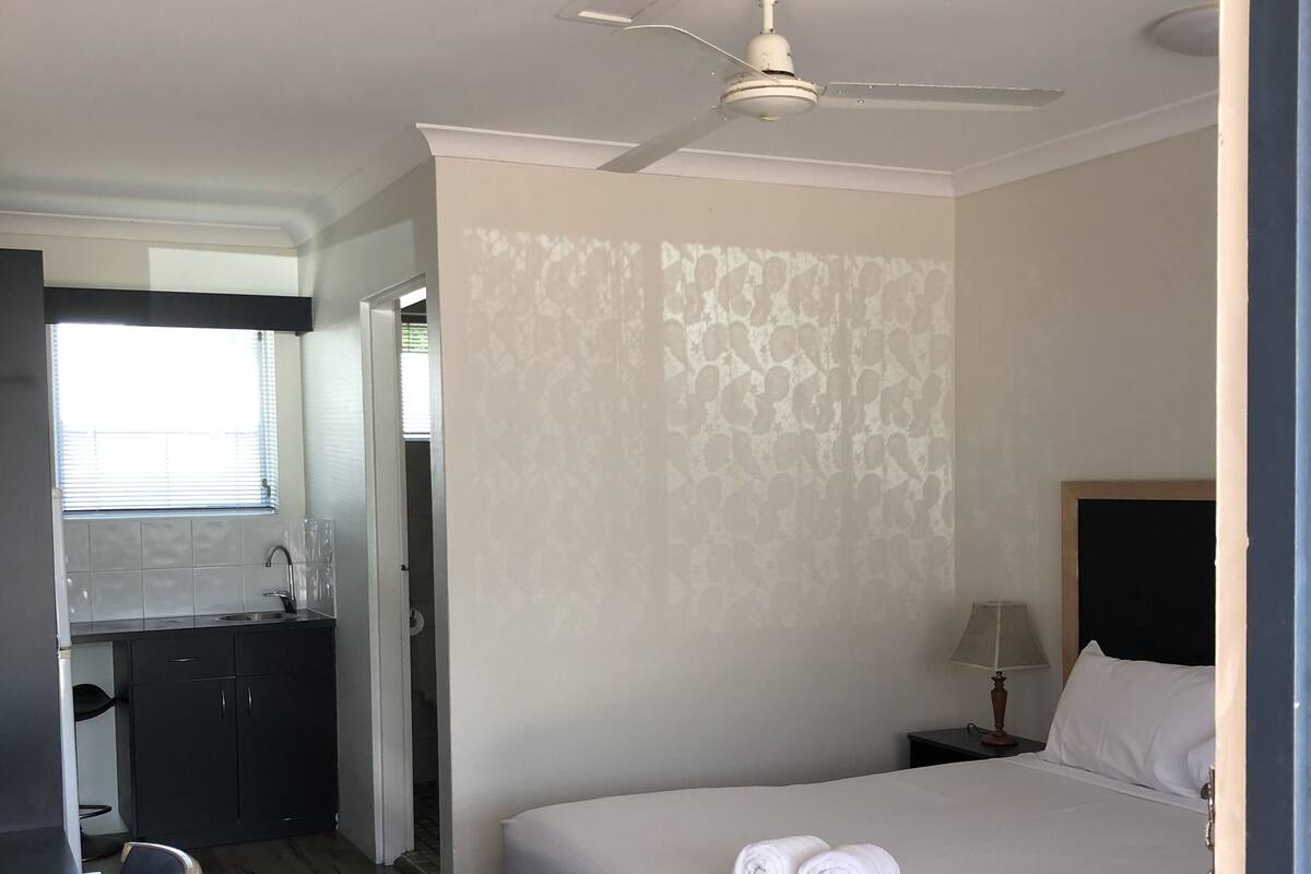 Quarto de hotel com cama, escrivaninha e cadeira. Há um ventilador de teto sobre a cama.