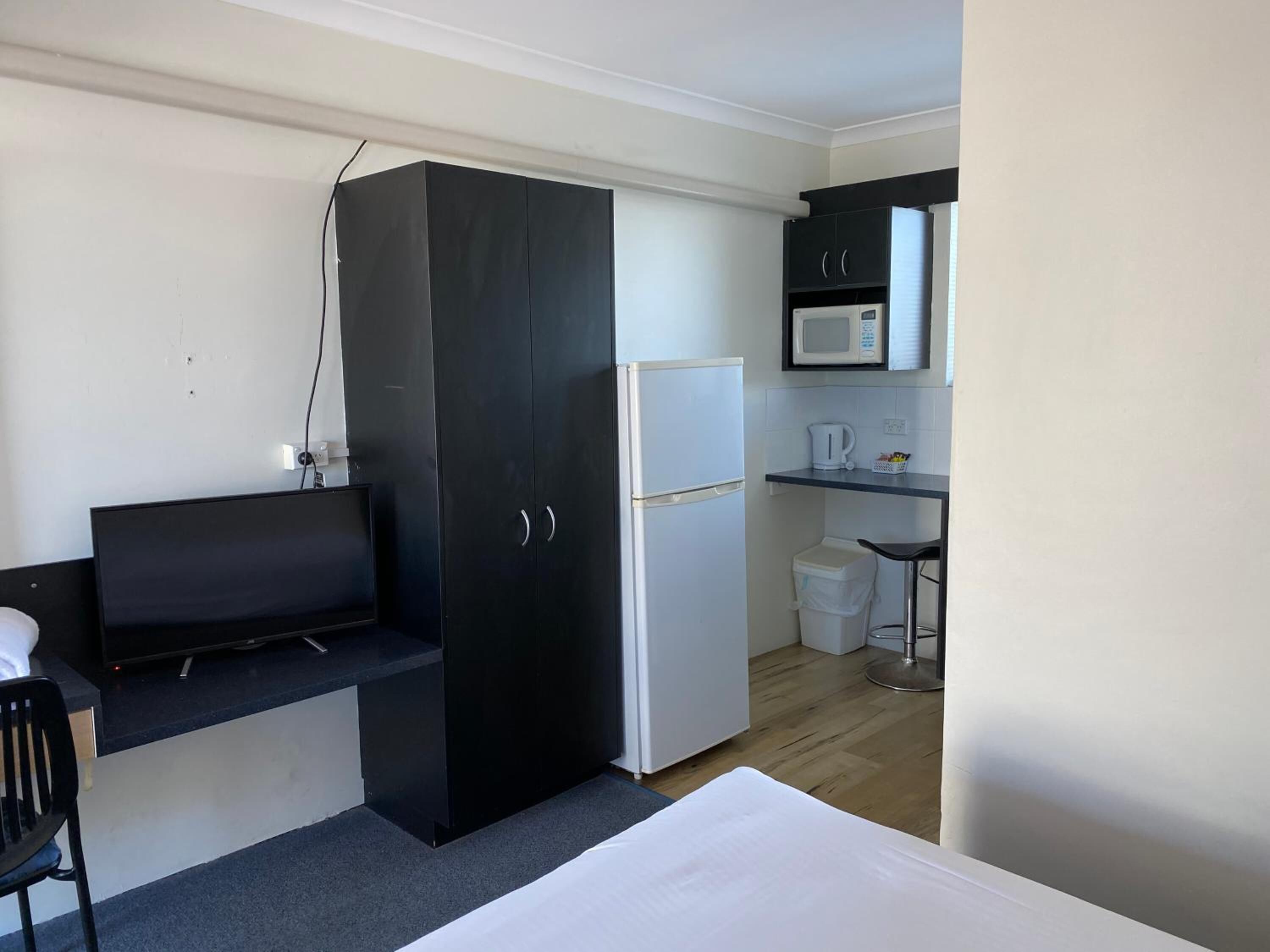 A compact room with a bed, a black wardrobe, a white refrigerator, a microwave, and a small kitchenette area.