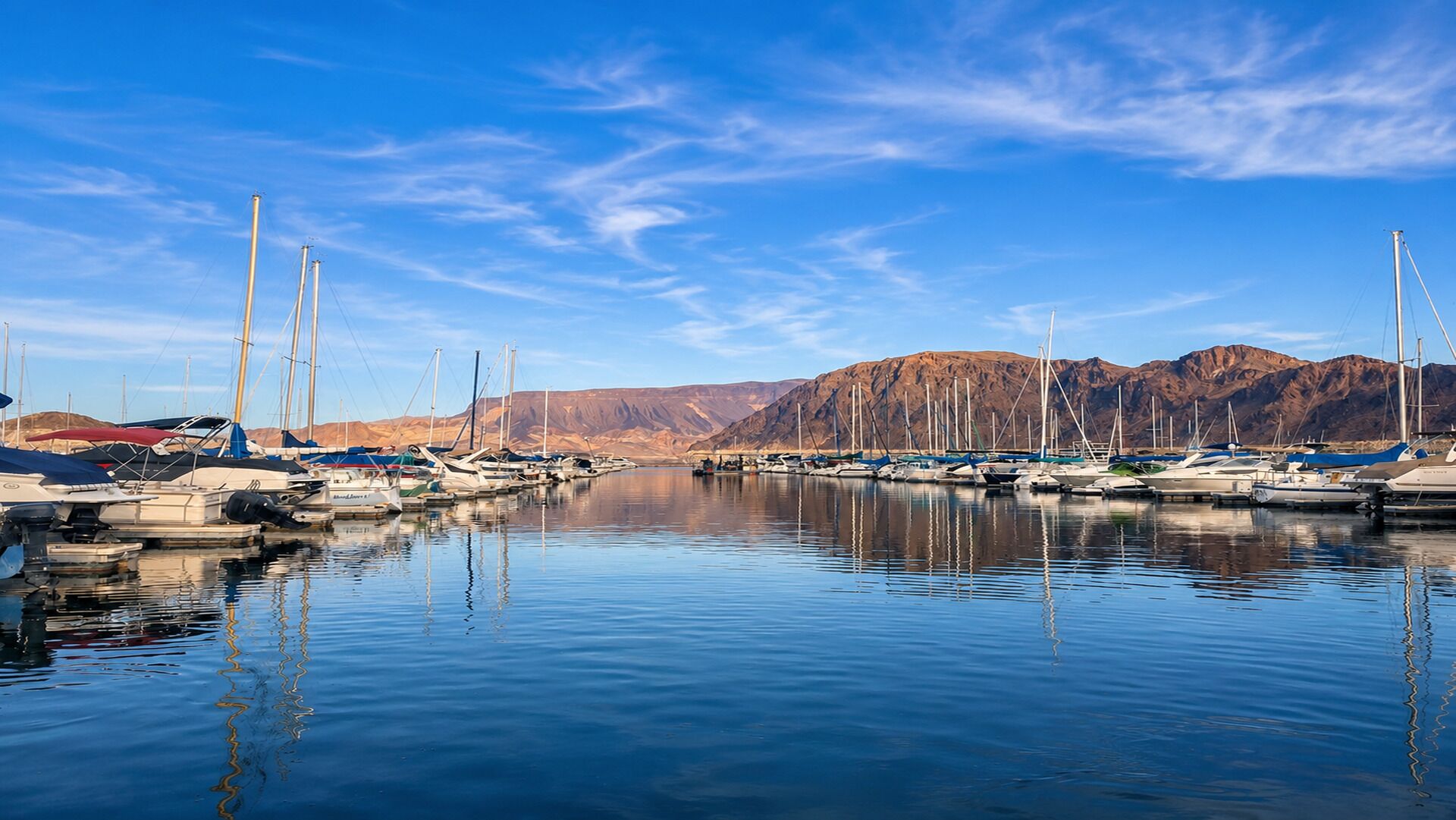 A marina with boats docked, calm water, and mountains in the background.