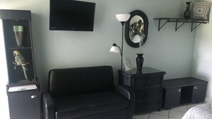 Une pièce avec un canapé noir, un téléviseur fixé au mur, une lampe, un miroir et une petite étagère ornée d’objets décoratifs.