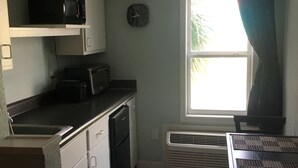 Une cuisine compacte avec des meubles blancs, un micro-ondes noir, un grille-pain et un réfrigérateur noir. On y trouve une fenêtre avec des rideaux, une horloge murale et une grille d’aération blanche.