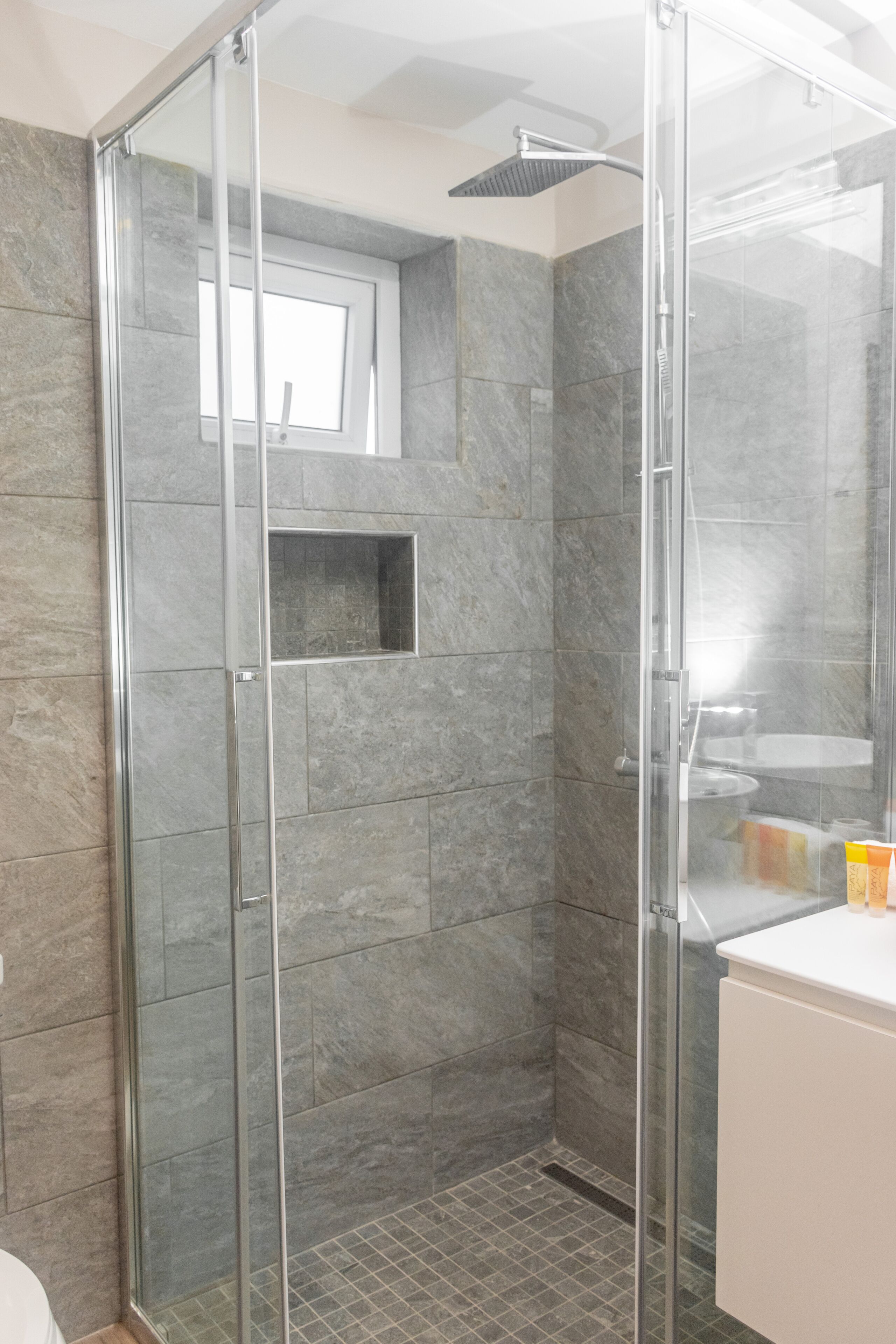 Un baño moderno con ducha envuelta en vidrio, baldosas de mármol gris y una pequeña ventana.