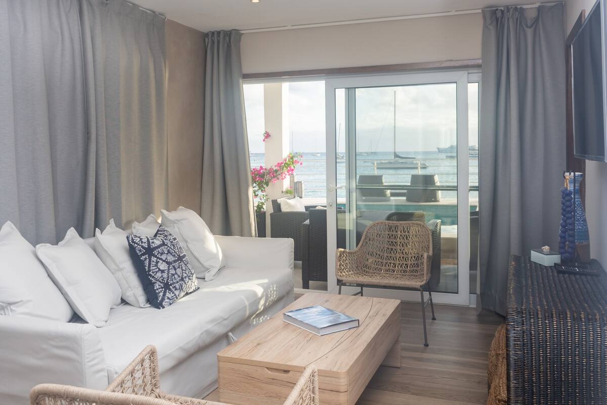 A modern living room with a sofa, a coffee table, and a wicker chair. There is a glass door leading to a balcony with a view of the sea and boats.