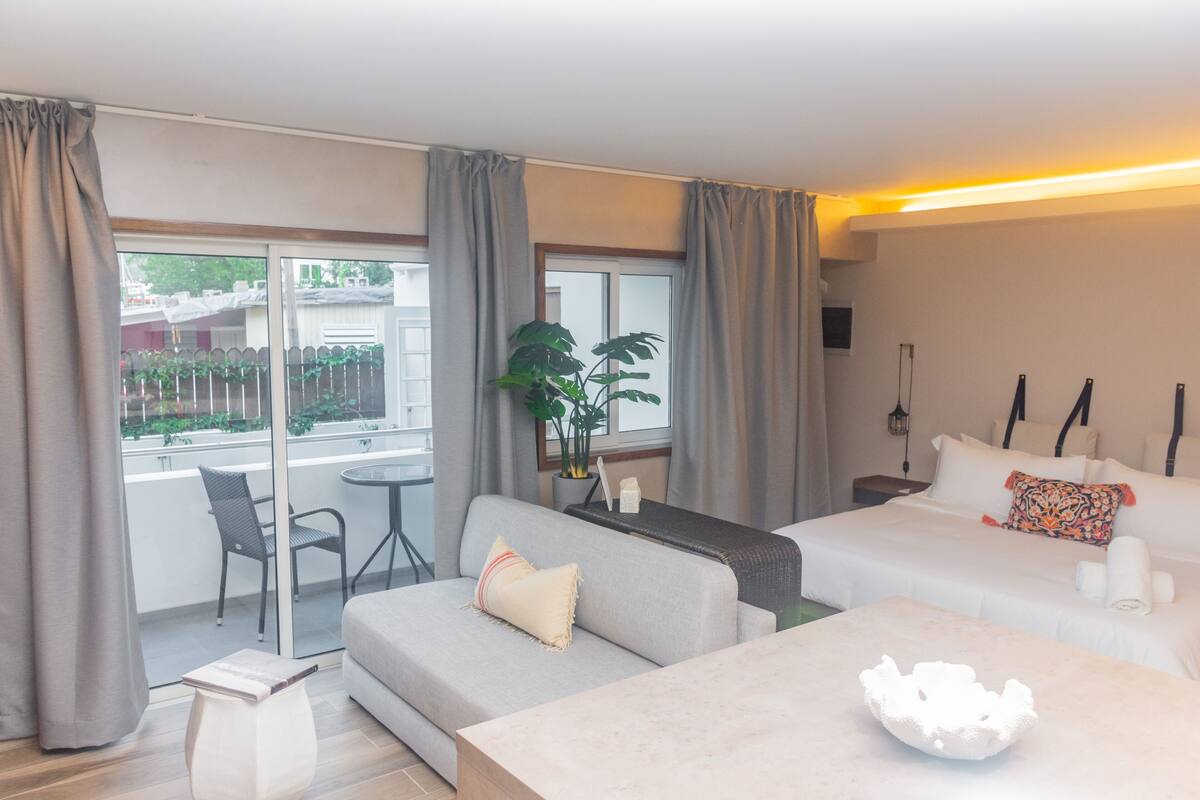 A hotel room with a sofa, a bed, a small table, a plant, and a view of the outside.