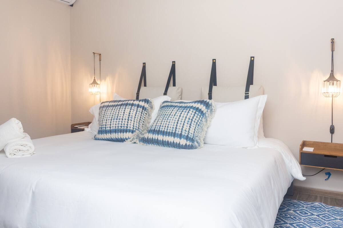 A neatly made bed with white bedding and two blue and white patterned pillows. There are two bedside tables, one with a lamp and the other with a small wooden tray.