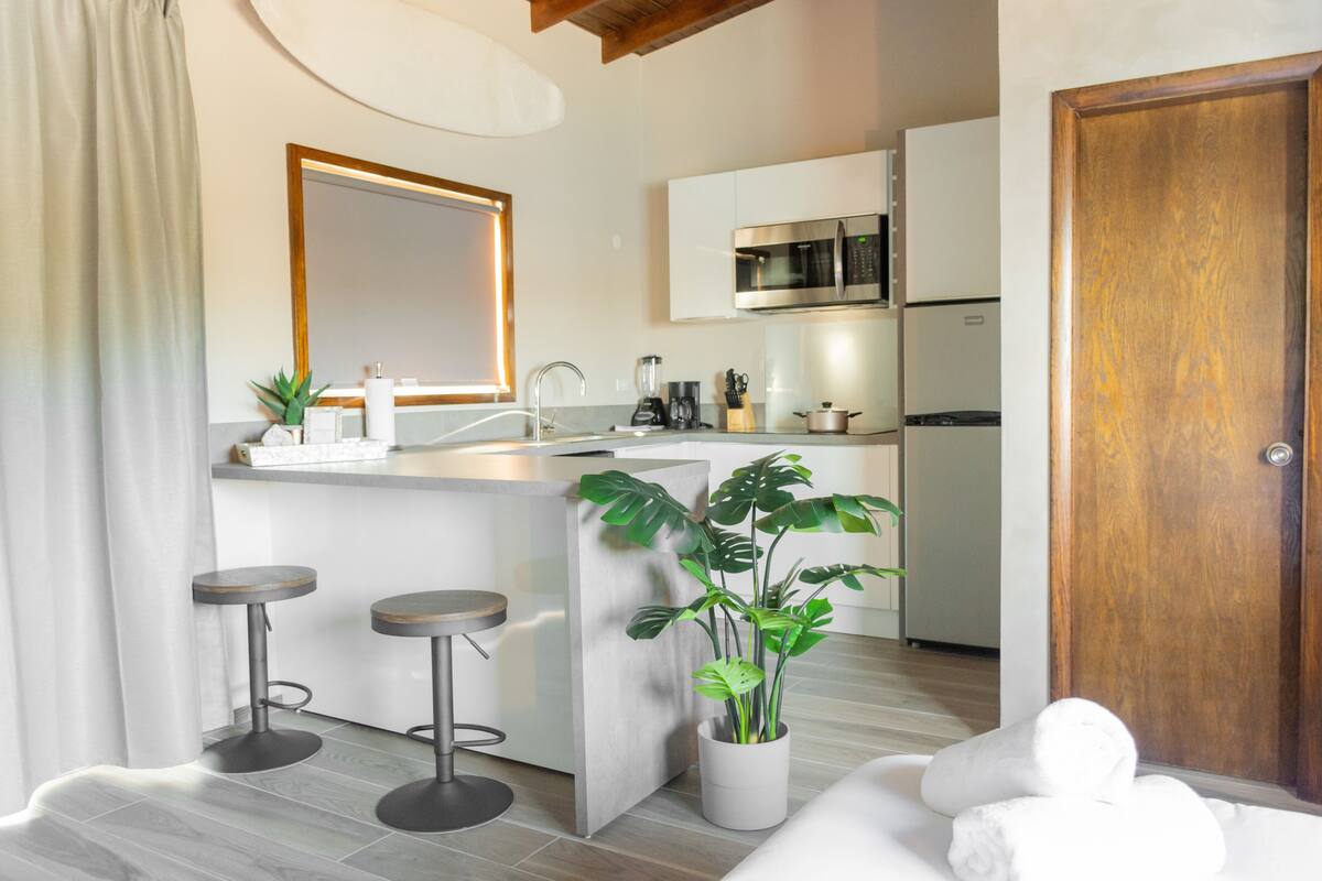 A modern kitchen with a central island, bar stools, a potted plant, and a wooden door.
