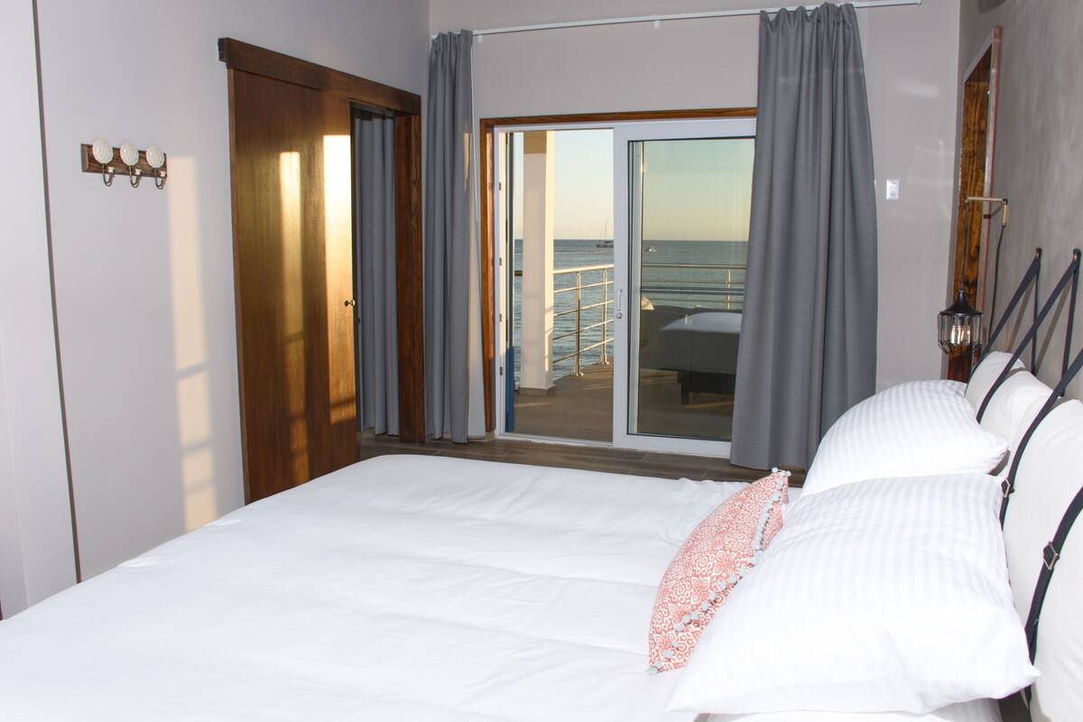 A hotel room with a large bed, a view of the sea through a sliding door, and a balcony with a railing.